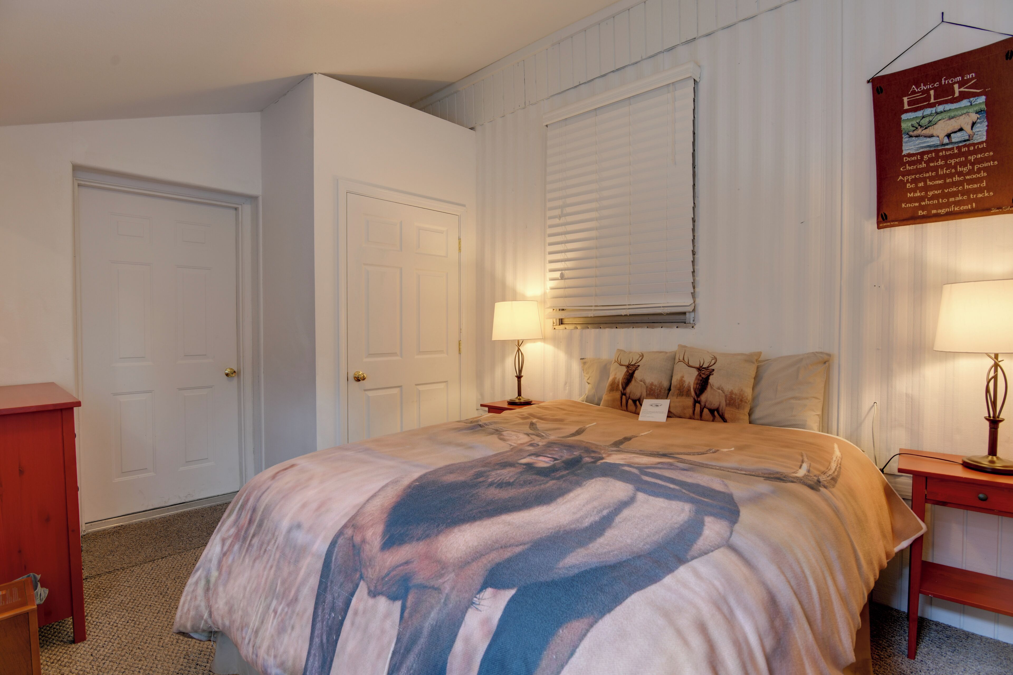 A bedroom with a bed, two bedside tables, a nightstand, a lamp, a door, and a window with blinds.