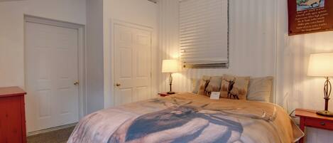 A bedroom with a bed, two bedside tables, a nightstand, a lamp, a door, and a window with blinds.