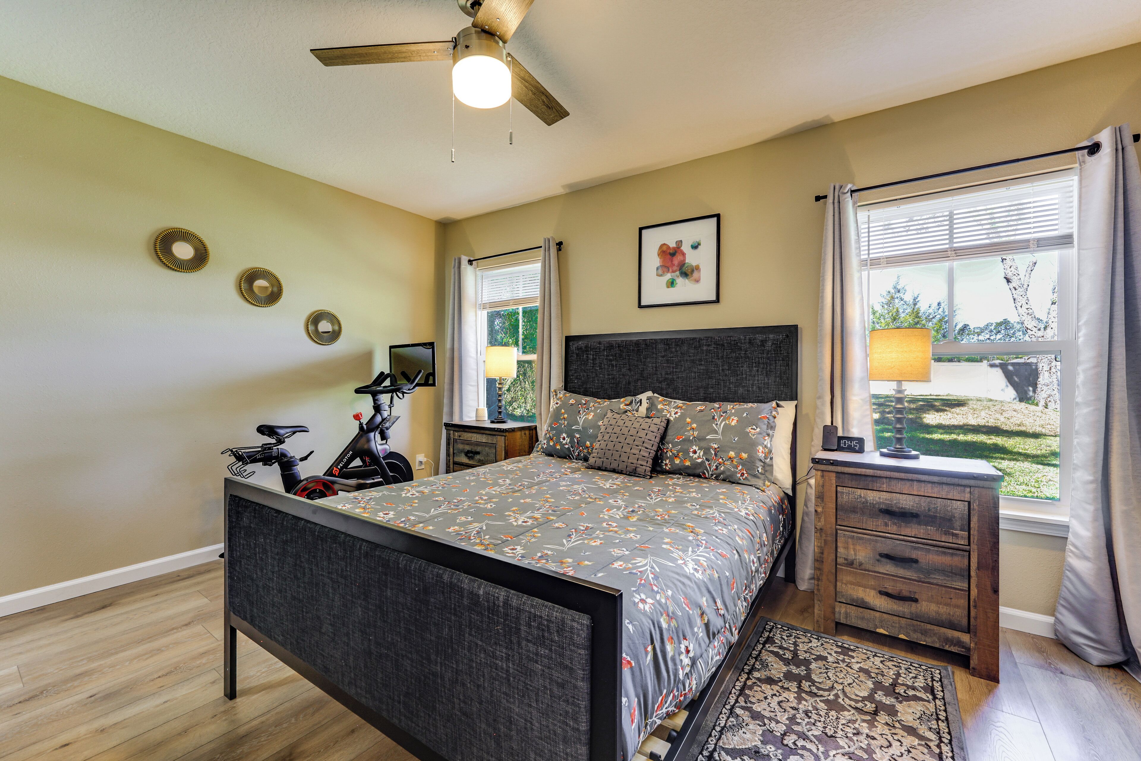 Un dormitorio con cama, ventilador de techo, ventana con cortinas, mesita de noche y una bicicleta.