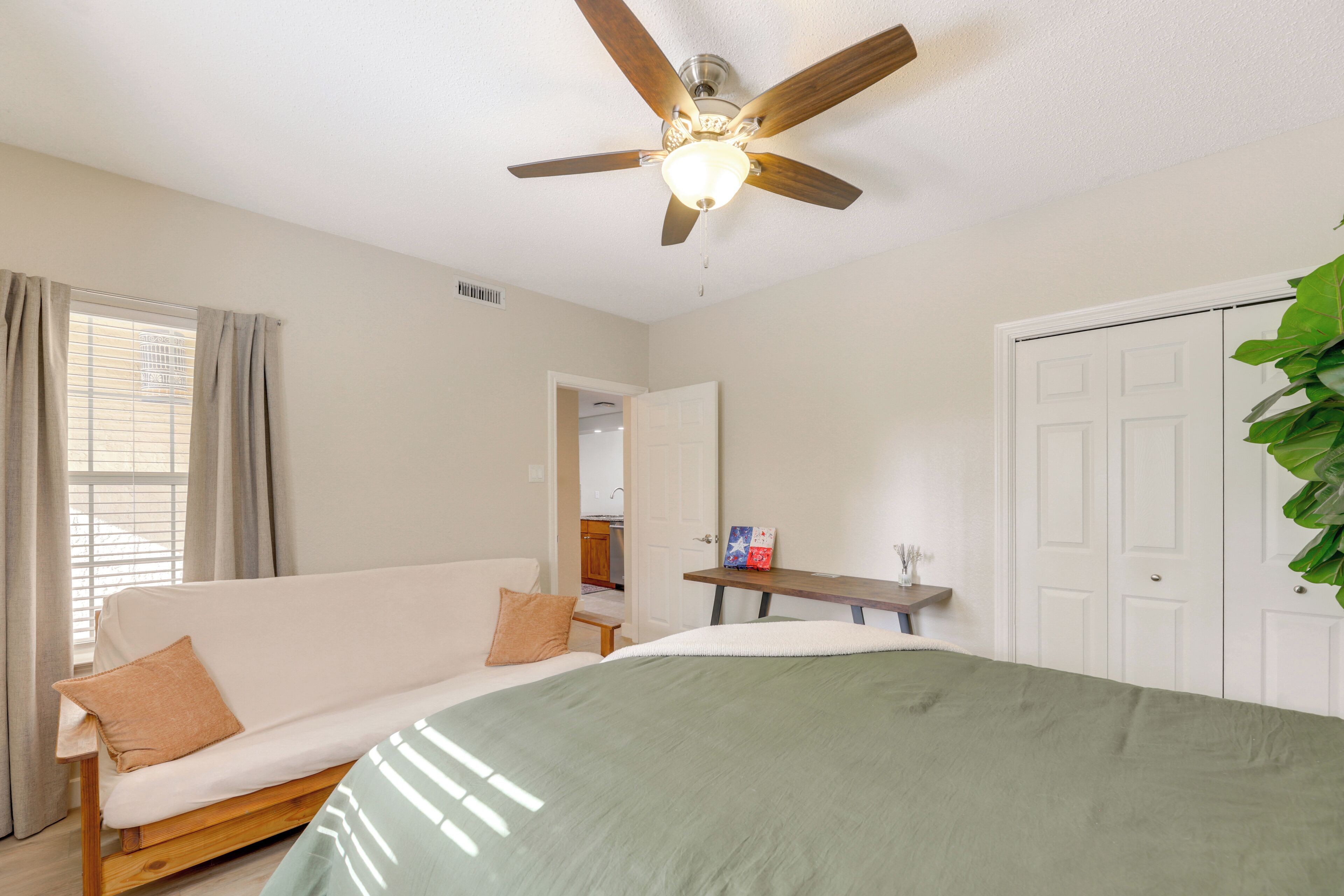 A bedroom with a bed, a ceiling fan, a window with curtains, a door, and a plant.