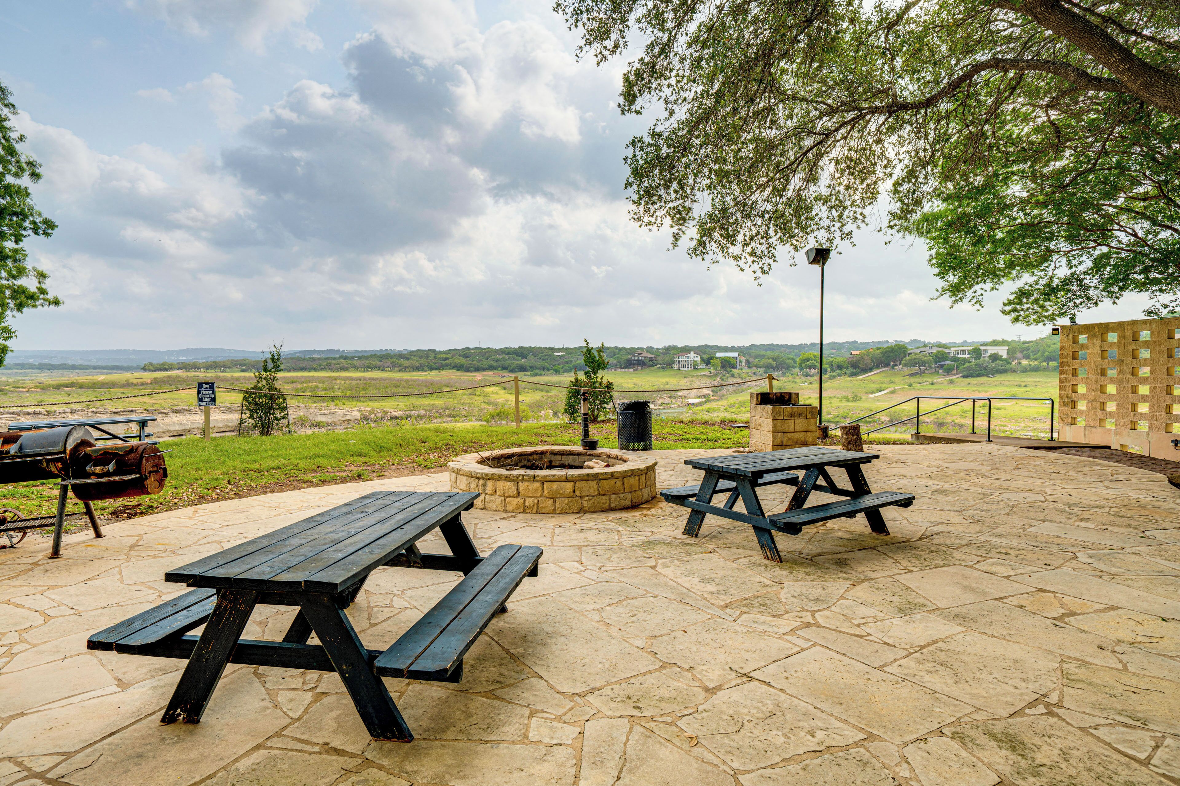 An outdoor patio with picnic tables and a fire pit, surrounded by stone flooring and a view of the countryside.