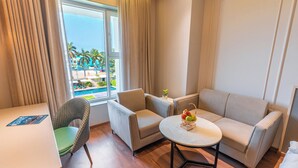 A hotel room with a sofa, armchair, small table, and a view of a pool and palm trees. - Ranveer the Garden Resort (Haldwani)