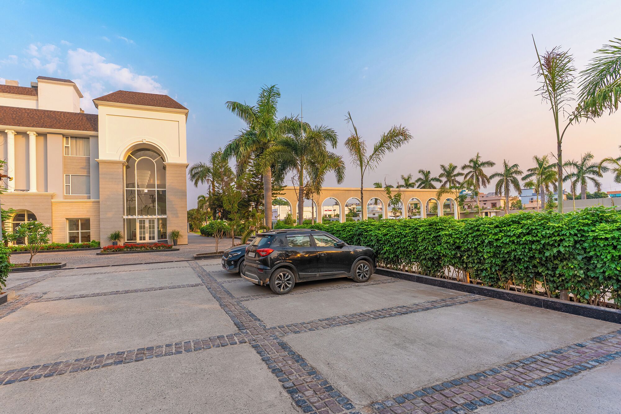 A building with a large arched entrance, a paved driveway, and a parking area with two cars.