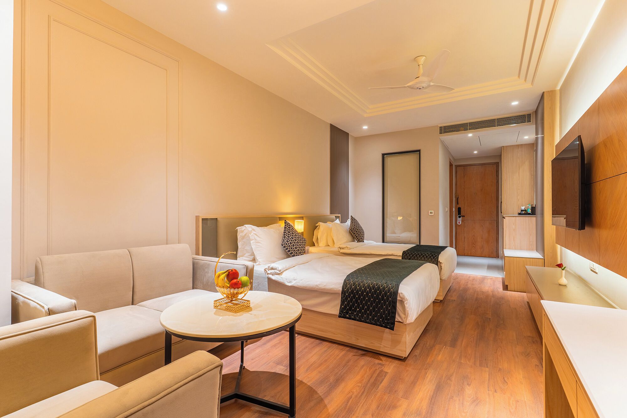 Una habitación de hotel con sofá, una mesita redonda con frutero, cama con cabecera, mesita de noche y televisor montado en la pared.