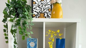 A white shelving unit with various items including a potted plant, framed artwork, a vase with flowers, a blue box, and a book titled 'Great Journeys'.