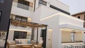 Een modern, tweelaags gebouw met een patio, voorzien van rieten meubels, een pergola en een bar met krukken.