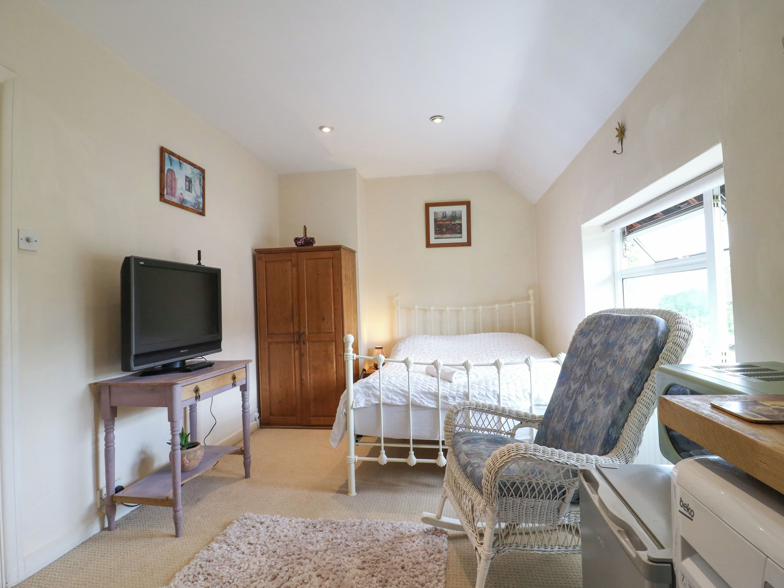 A bedroom with a bed, a television on a wooden table, a wicker chair, a wardrobe, and a window.