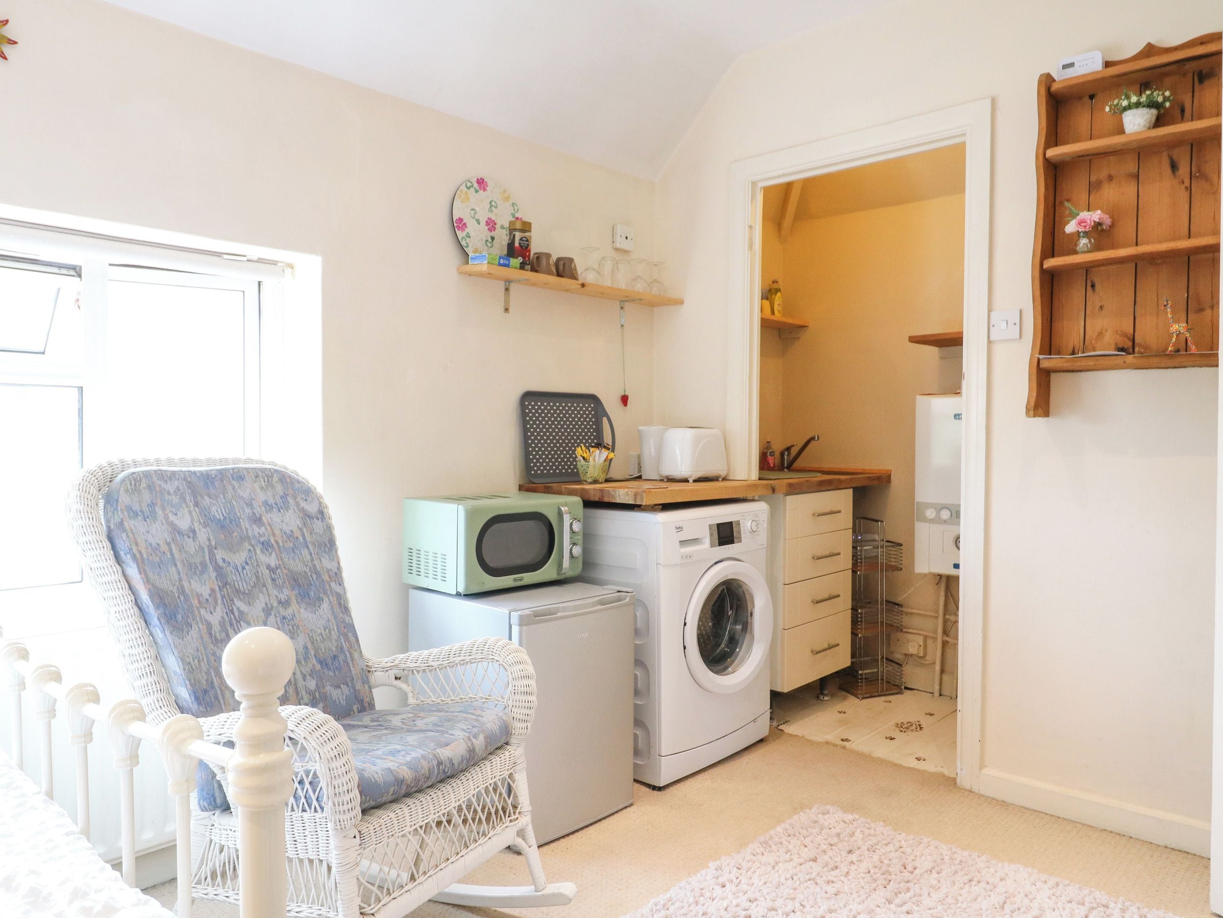 A compact living space with a washing machine, a microwave, a wicker chair, and a small kitchenette.