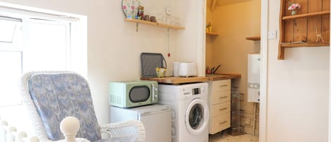 A compact living space with a washing machine, a microwave, a wicker chair, and a small kitchenette.