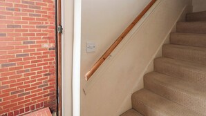 Eine Treppe mit Holzwah und beigefarbenem Teppich, die zu einer Tür an einer Ziegelwand führt, mit Lichtschalter an der Wand.