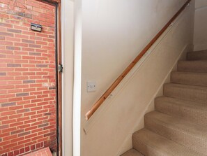 A staircase with a wooden handrail and beige carpeted steps, leading to a door with a brick wall and a light switch on the wall. - Daisy Cottage (Dorking)