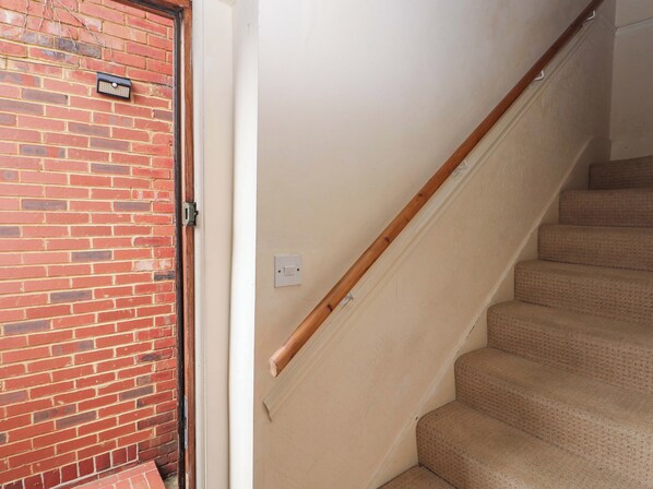 A staircase with a wooden handrail and beige carpeted steps, leading to a door with a brick wall and a light switch on the wall.