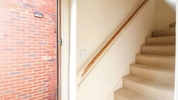 A staircase with beige carpeted steps and a wooden handrail, leading to a door with a red brick wall.