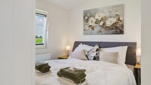 A neatly made bed with folded towels, a painting of flowers on the wall, and a window with a view of greenery.