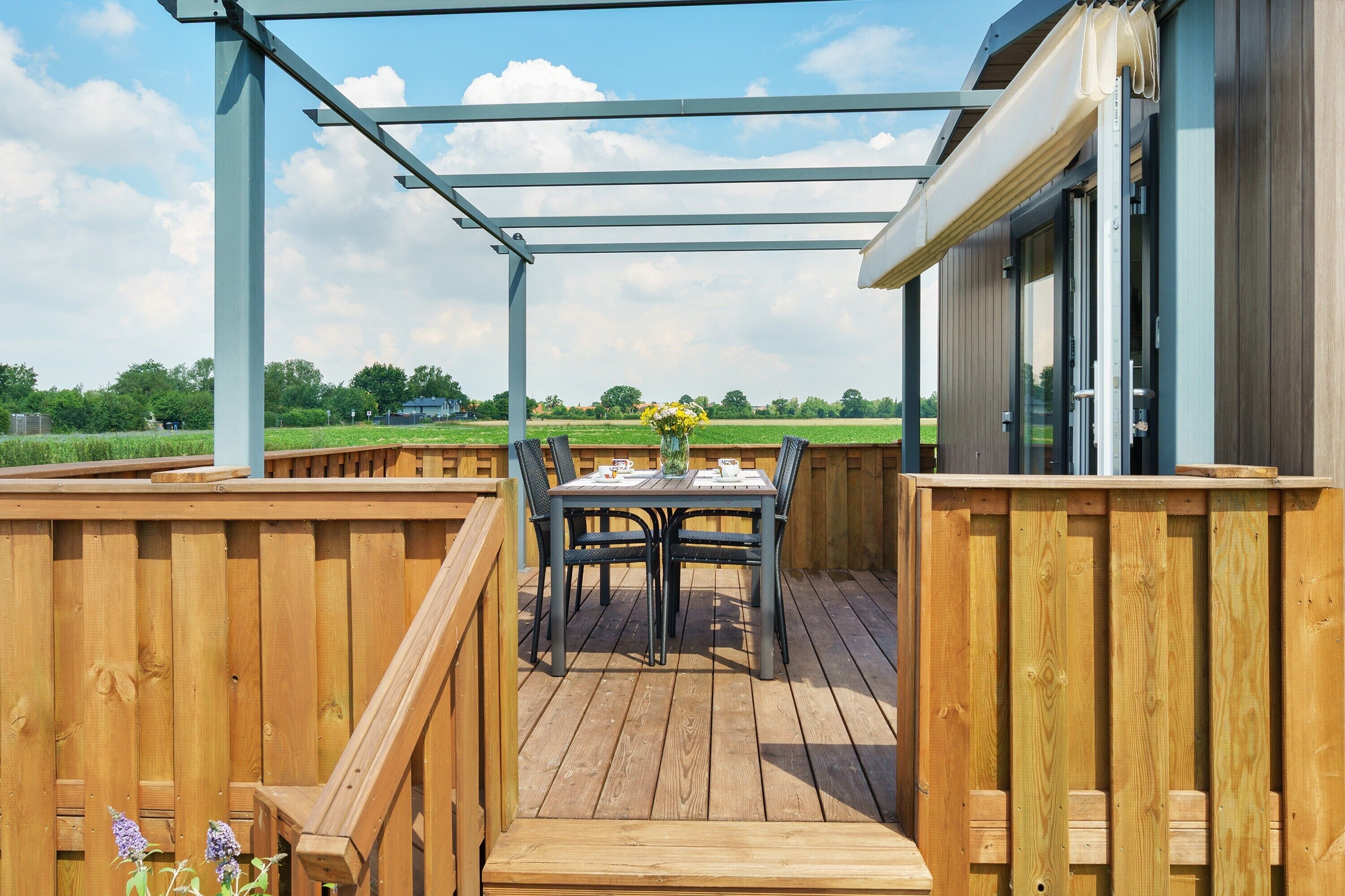 Eine Holzterrasse mit Tisch und Stühlen, einer Pergola und Blick auf ein grünes Feld.