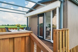 A wooden deck with a table and chairs, a sliding glass door leading to an indoor area with a sofa and a plant. - Chalet 'Rotkäppchen' with Nature Views by the Lake (Dörverden)