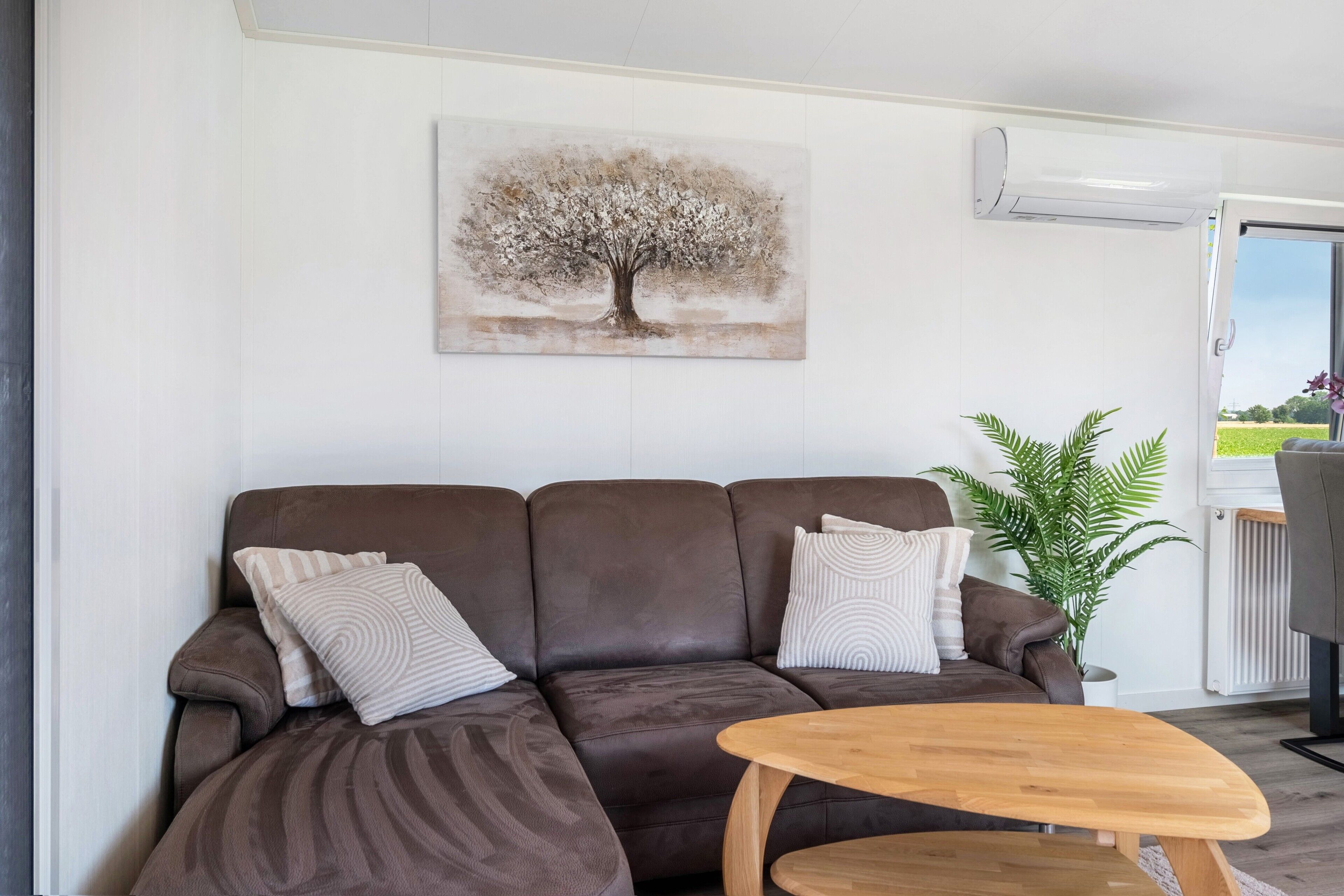 A modern living room with a brown sofa, patterned cushions, a wooden coffee table, and a painting of a tree.