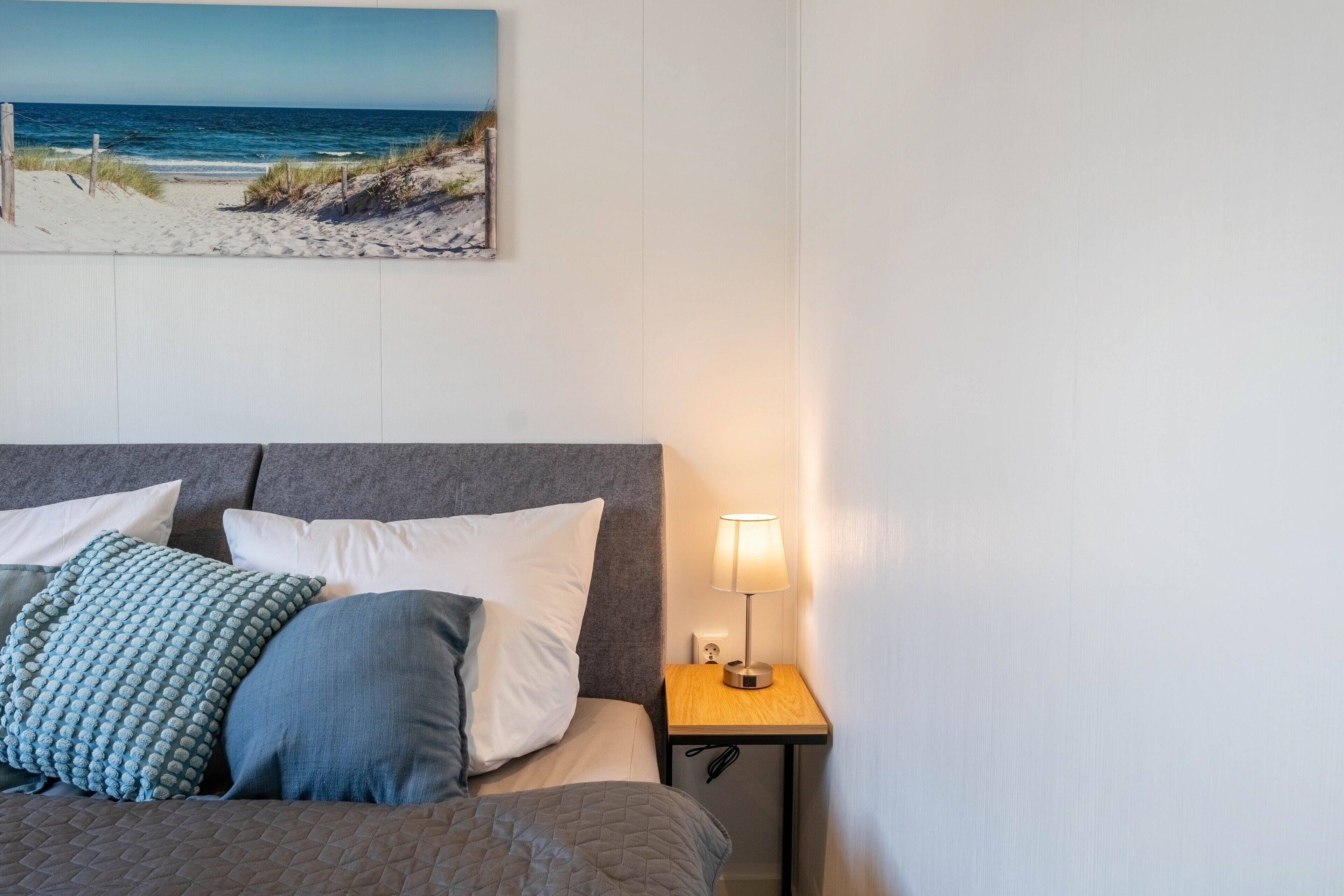 A neatly made bed with pillows, a bedside lamp, and a painting of a beach scene on the wall.