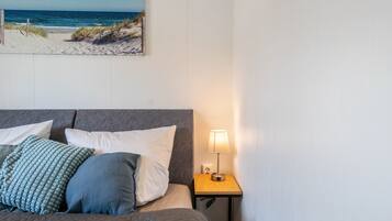 A neatly made bed with pillows, a bedside lamp, and a painting of a beach scene on the wall.