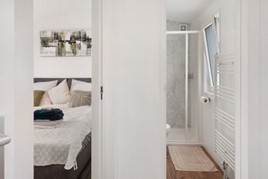 A bedroom with a bed, a bedside table, a shower, and a door leading to another room.