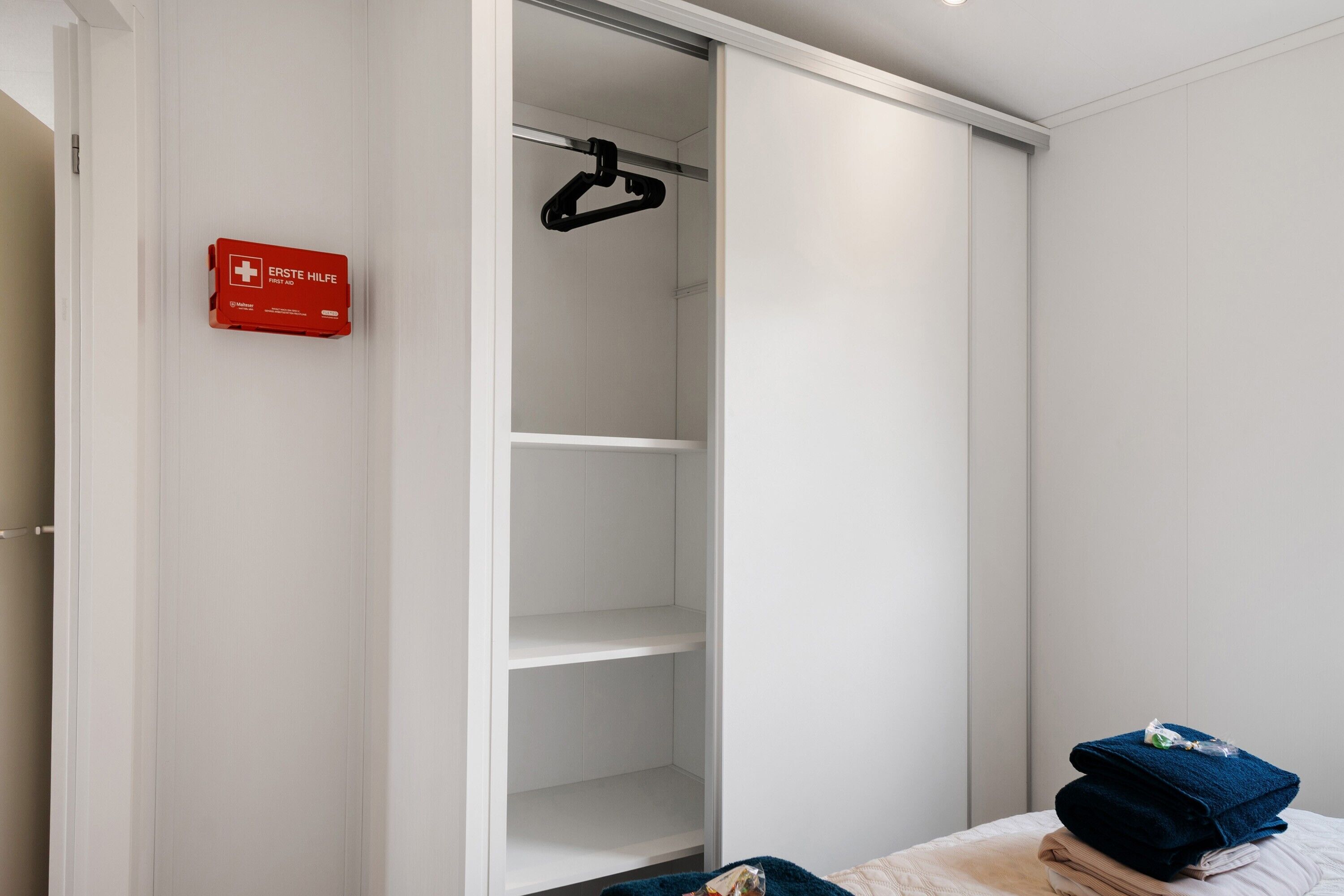 A bedroom with a white wardrobe, a red emergency aid sign, and a bed with folded towels.