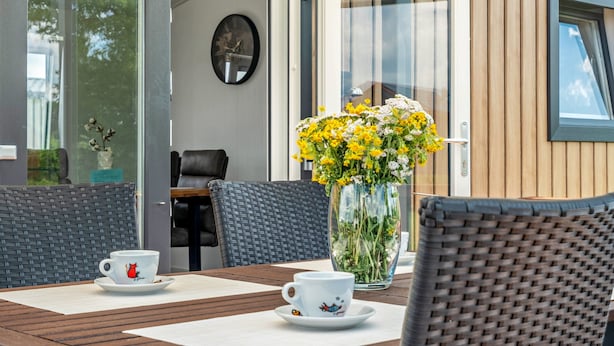A dining area with wicker chairs, a table with a placemat, a vase with yellow flowers, and a clock on the wall.