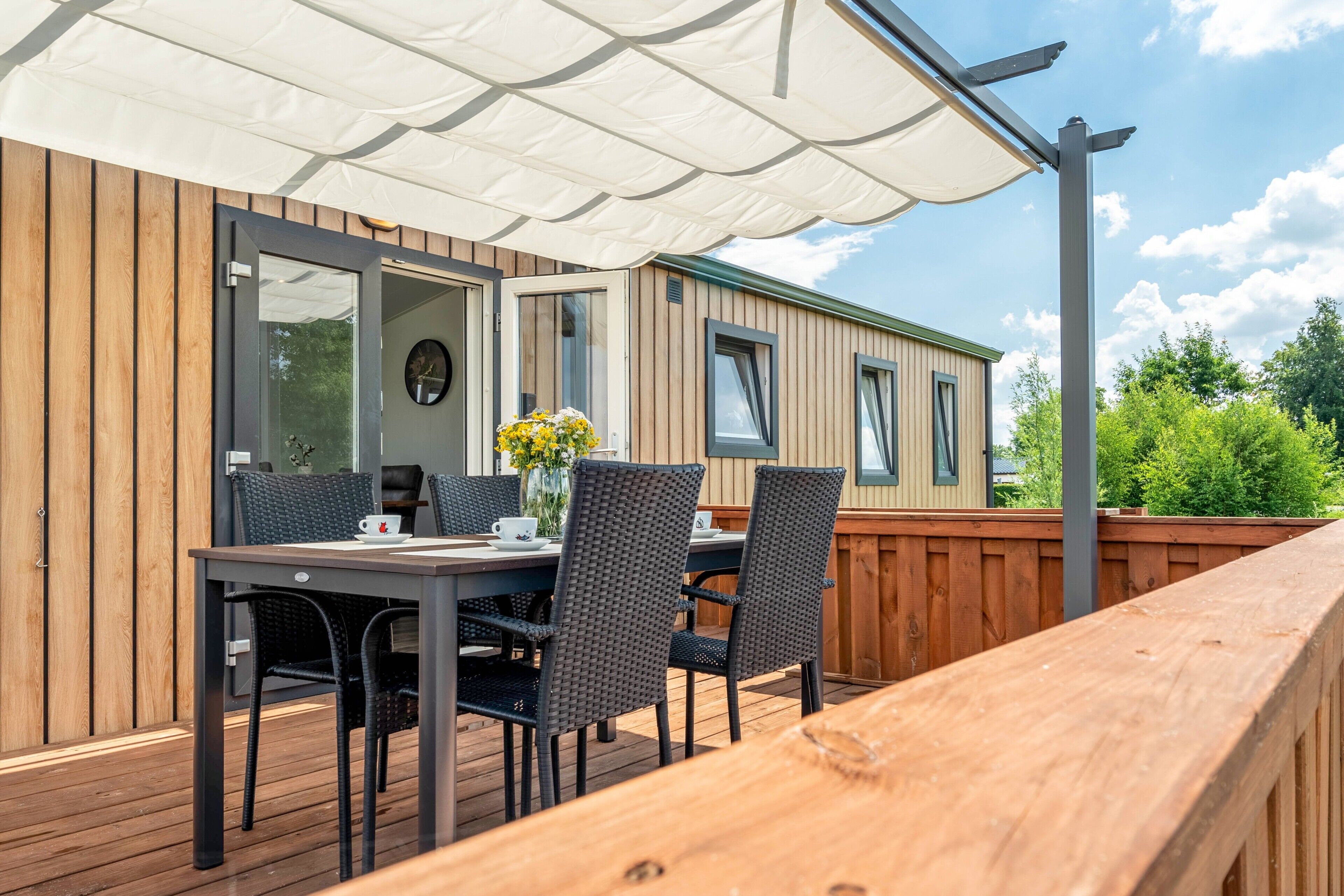 Um deck de madeira com mesa e cadeiras, um toldo branco e vista para uma casa móvel.