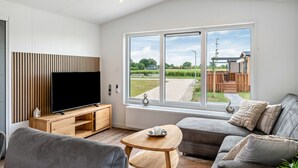 A modern living room with a flat-screen TV, wooden furniture, a grey sofa, and a large window offering a view of the outdoors.