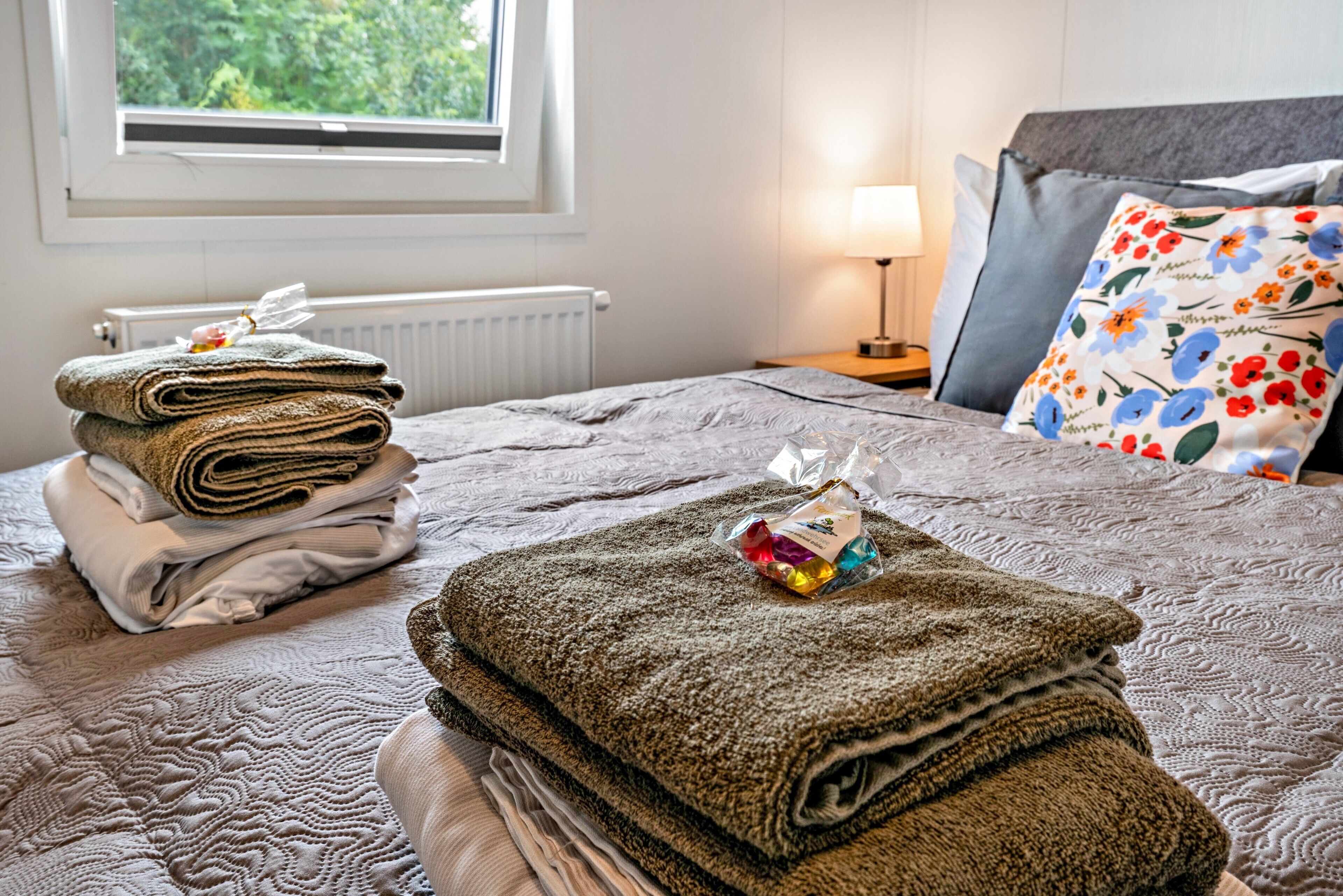 A neatly made bed with folded towels, a lamp, and a window with a view of greenery.