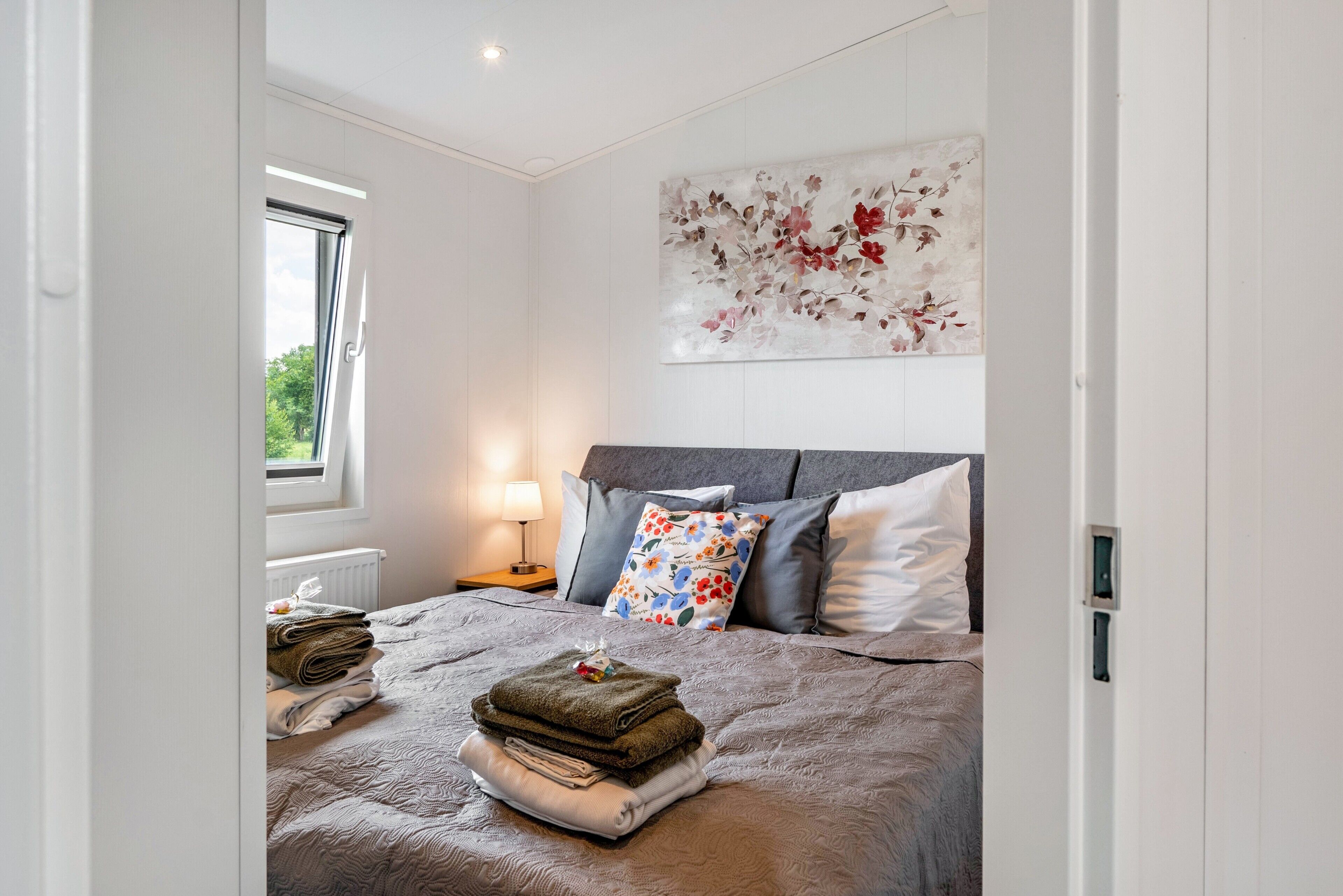 Ein Schlafzimmer mit einem Bett, Kissen, einem Nachttisch, einer Lampe, einem Fenster und einem floralen Kunstwerk an der Wand.