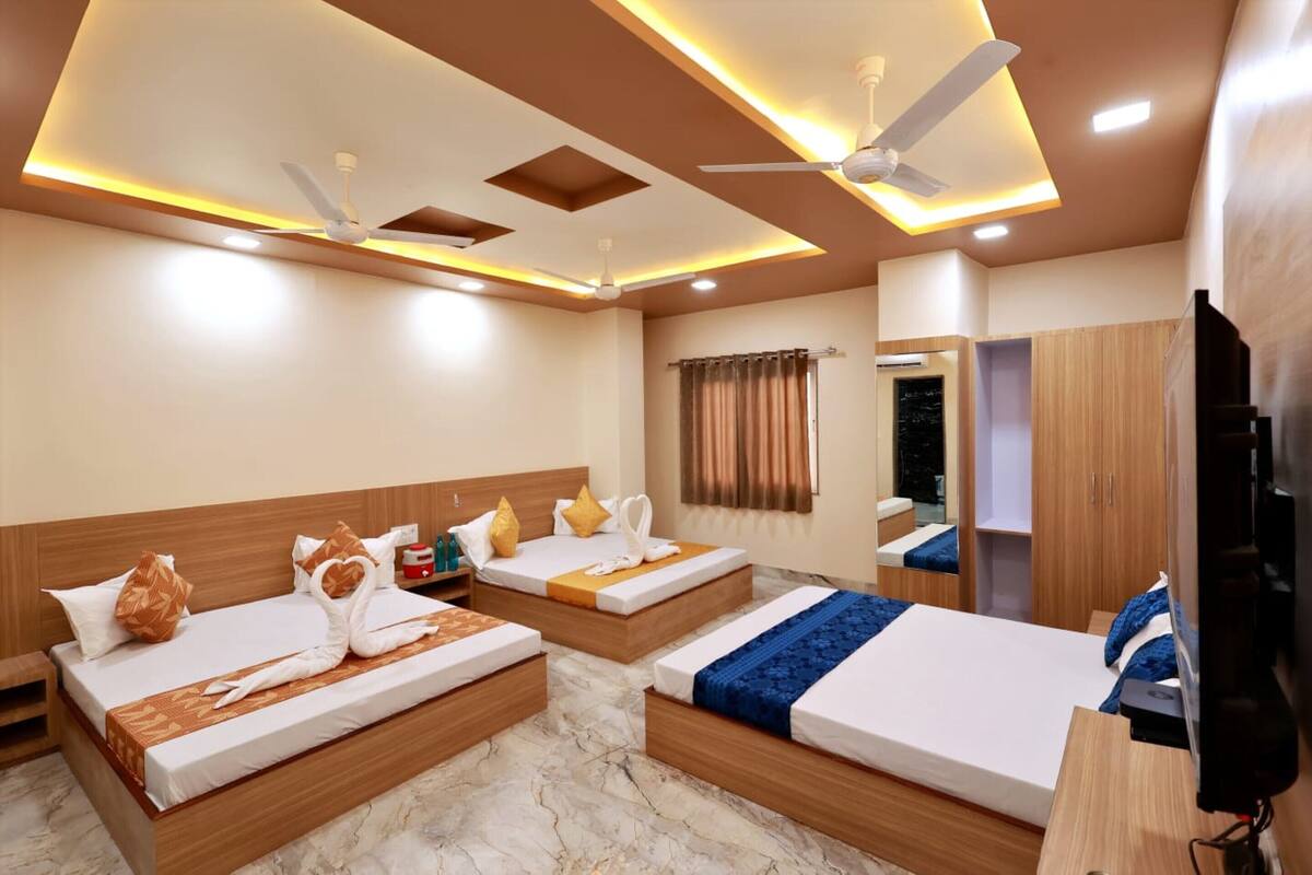 Una habitación de hotel con tres camas, ventiladores de techo, iluminación empotrada, un televisor y un armario.