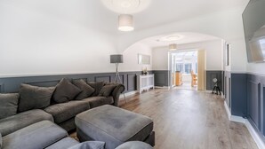 Ein Wohnzimmer mit einem grauen Ecksofa, einer Stehlampe und einem Essbereich im Hintergrund.