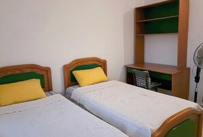 Ein Zimmer mit zwei Einzelbetten, jeweils mit einem hölzernen Kopfteil und einem grünen Kissen. Es gibt einen kleinen Schreibtisch mit Stuhl und ein grünes Regal darüber.