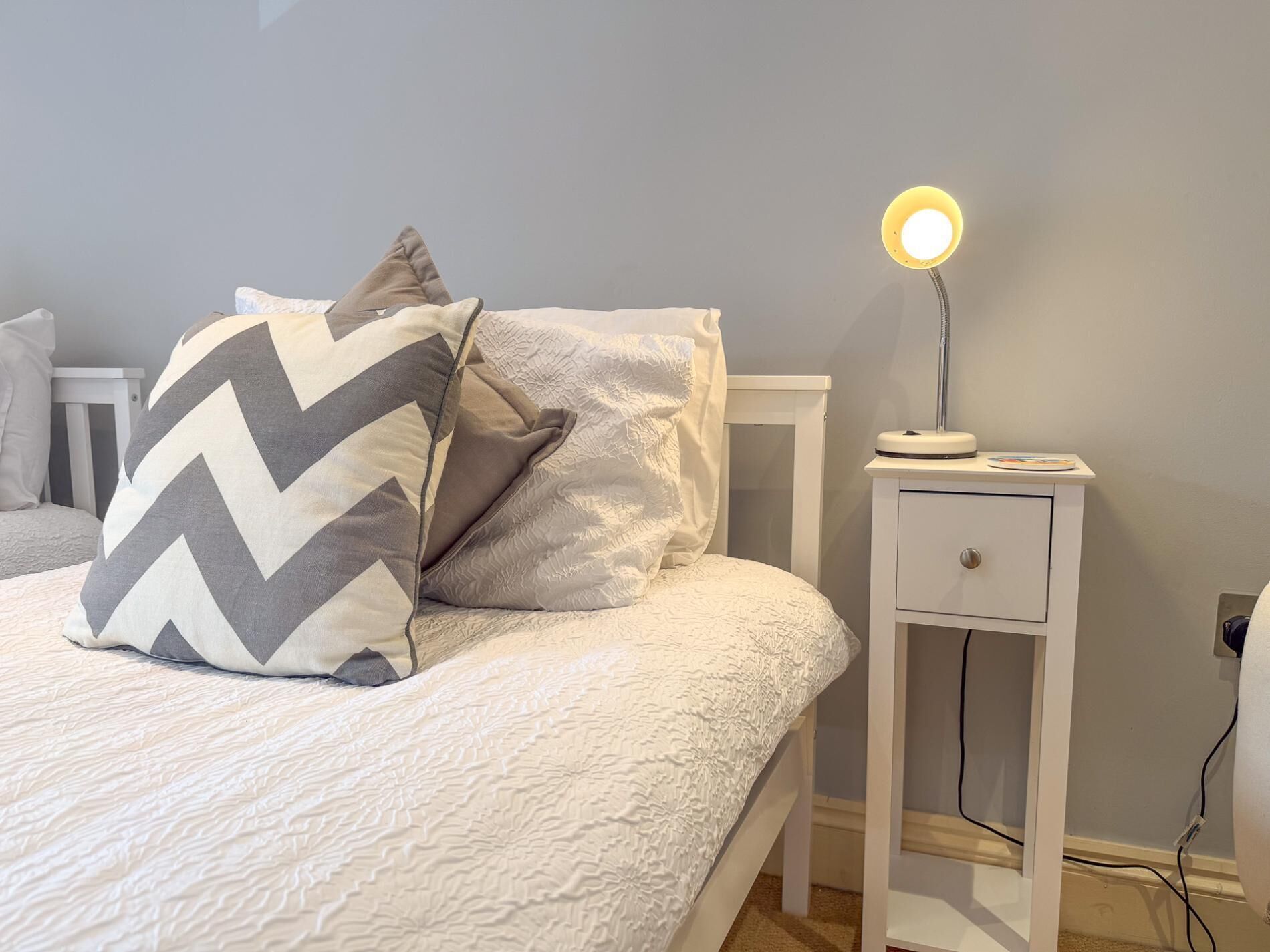 Una cama bien hecha con almohadones estampados, una lámpara de noche y una mesita de noche blanca con un cajón.