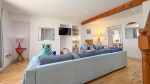Een ruime woonkamer met een grote bank, houten balken in het plafond, een flatscreen televisie en een trap die naar een hoger niveau leidt.