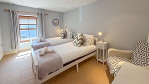 Een slaapkamer met twee bedden, een nachtkastje met een lamp, een stoel, een raam met gordijnen en een aan de muur hangend schilderij.