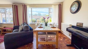A living room with a sofa, dining table, and piano. - Indigo Blue - 2 Bedroom Holiday Home - Manorbier (Tenby)