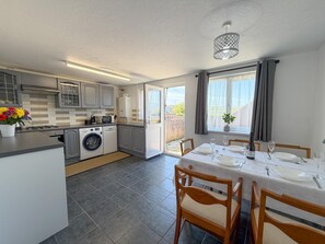 A modern kitchen with a built-in washing machine, a dining area with a table set for four, and a large window with curtains. - Pleasant View - 3 Bedroom Holiday Home - Pembroke (Pembroke)