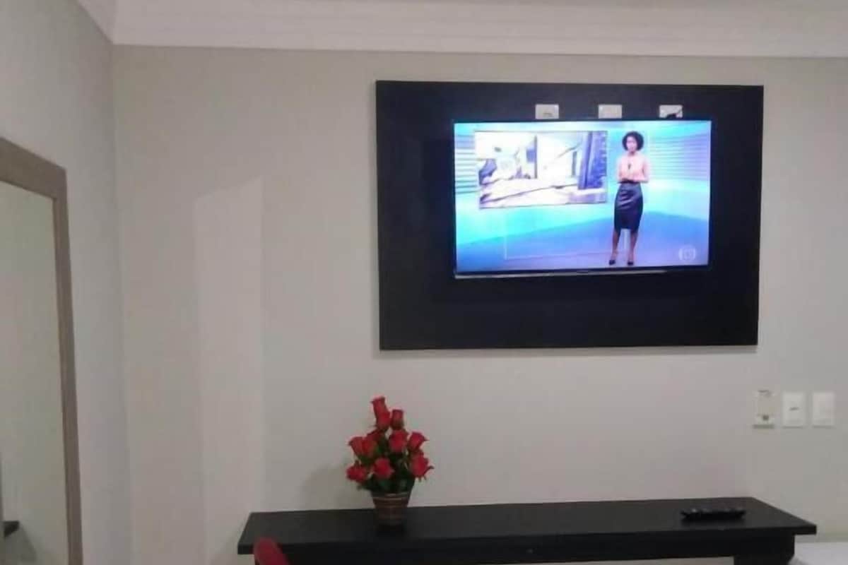 A room with a TV mounted on the wall, a red chair, a small table with a vase of flowers, and a white trash bin.