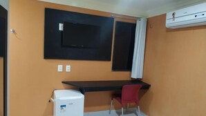 Una habitación pequeña con escritorio, una silla, un refrigerador blanco y un equipo de aire acondicionado.
