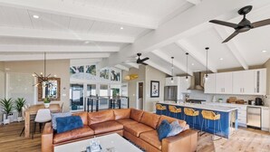 A spacious living area with a leather sofa, a coffee table, and a dining area with a table and chairs. There is a kitchen with white cabinets and a blue island. - Seacliff Ocean View Escape (Aptos)