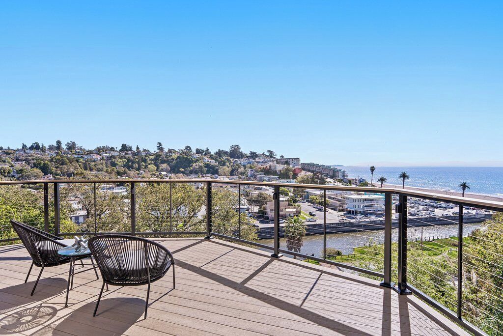 Eine Holzterrasse mit zwei Stühlen, einem Geländer und Blick auf Strand und Stadtlandschaft.