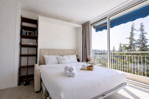 A hotel room with a large bed, a bookshelf, a balcony with a view of trees, and a window with curtains. - Studio Marina Mandelieu - Stylish Studio on the French Riviera (Mandelieu-la-Napoule)