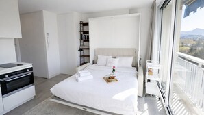 Moderni hotellihuone, jossa on sänky, pieni keittiö, parveke, jolta on näkymä, ja suuri ikkuna.