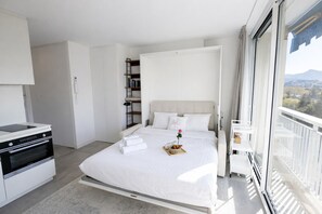 A modern hotel room with a bed, a kitchenette, a balcony with a view, and a large window. - Studio Marina Mandelieu - Stylish Studio on the French Riviera (Mandelieu-la-Napoule)