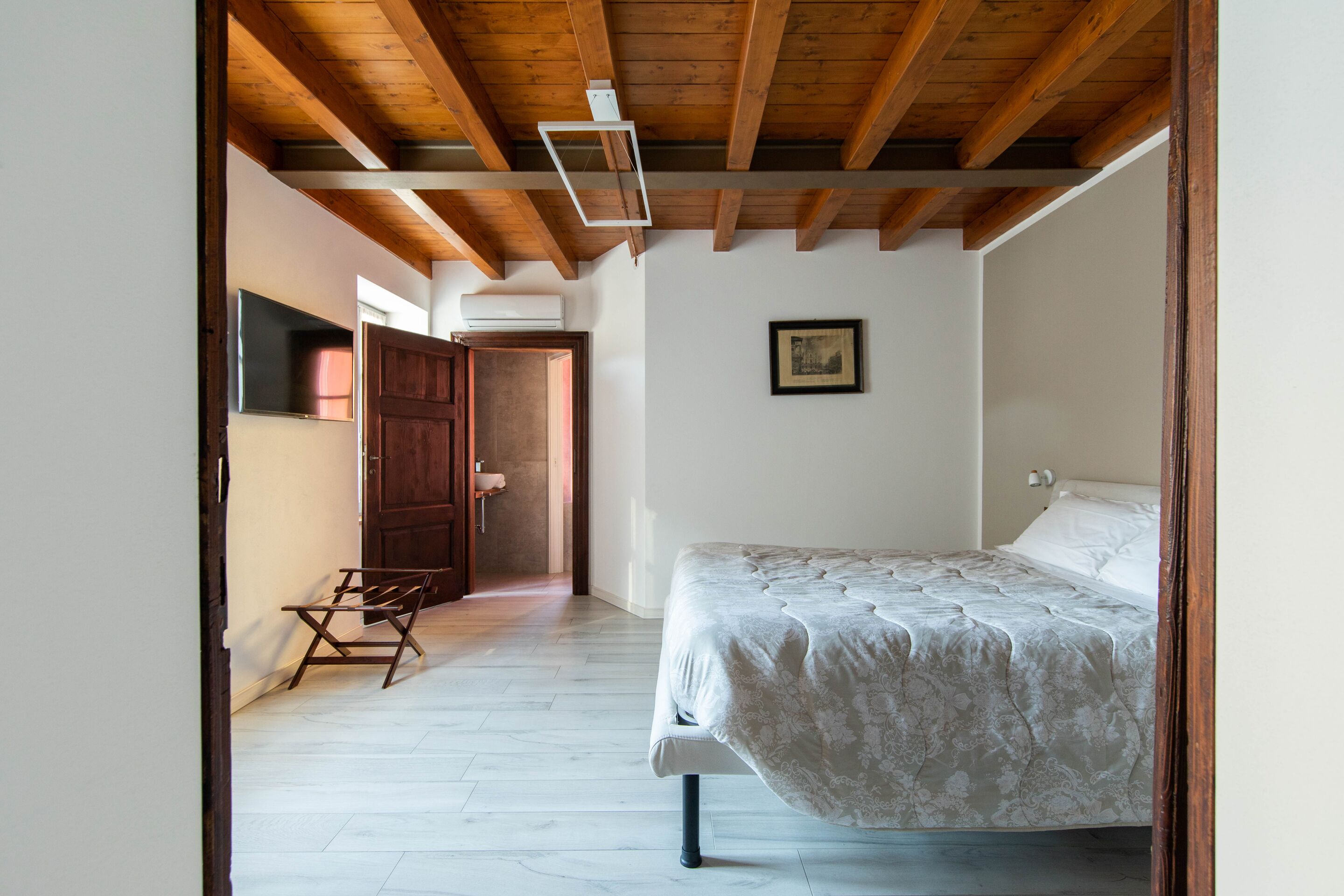 A bedroom with a bed, wooden ceiling beams, a framed picture on the wall, and a door leading to another room.