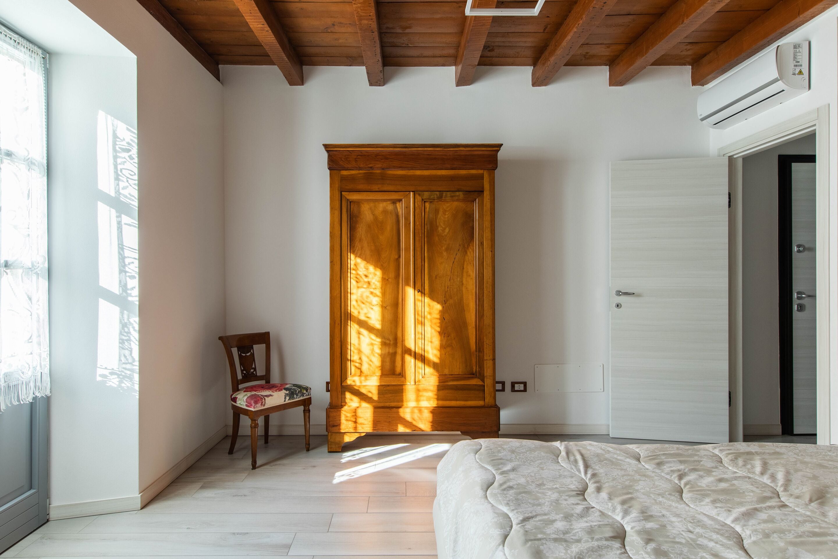 Una camera da letto con un letto, un'armadio in legno, una sedia e una finestra illuminata dal sole.