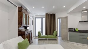 Ein modernes Hotelzimmer mit einem Bett, einem grünen Sofa, einem Holzschrank, einer Küchenzeile und einem durch die Tür einsehbaren Badezimmer.