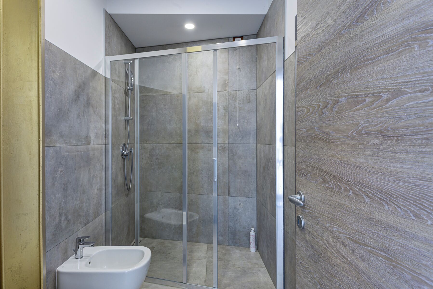 A modern bathroom with a glass shower enclosure, a bidet, and a wooden wall.