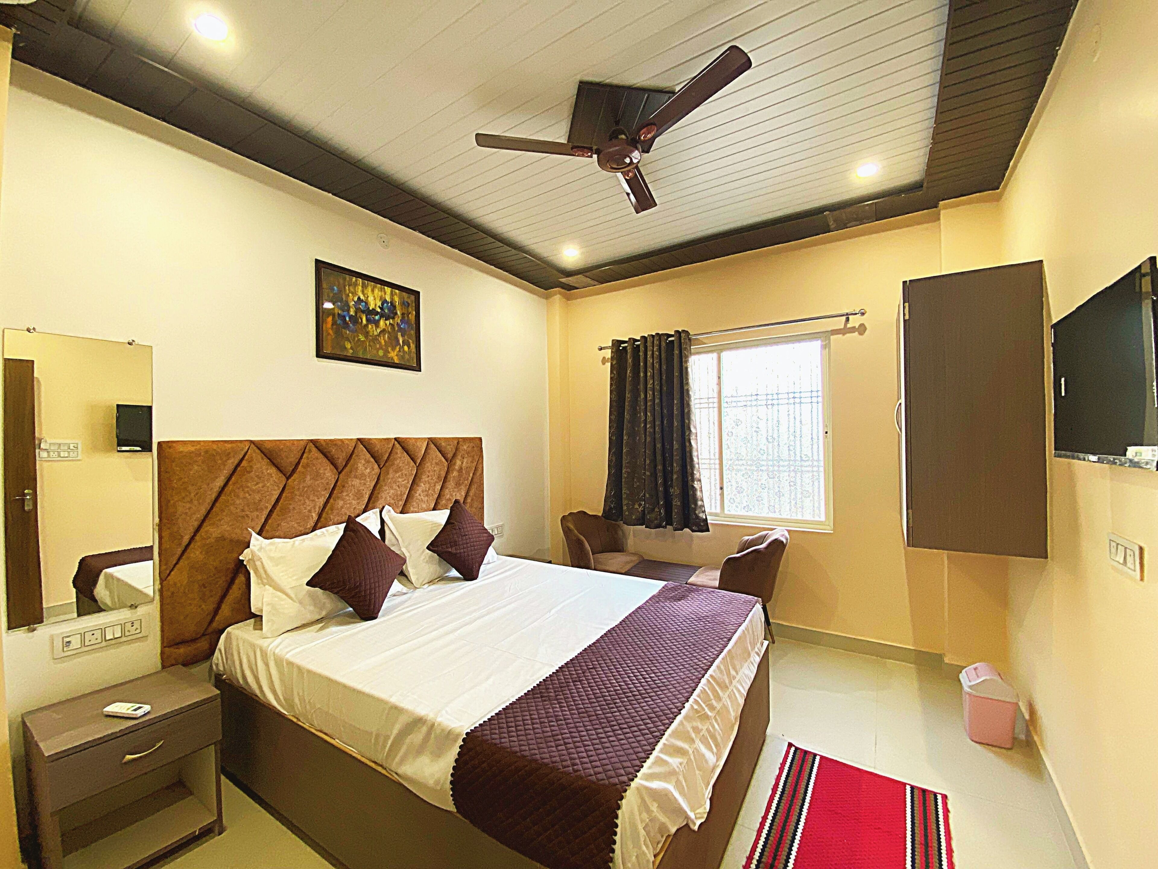 A hotel room with a bed, a desk, a chair, a ceiling fan, a TV, and a window with curtains.